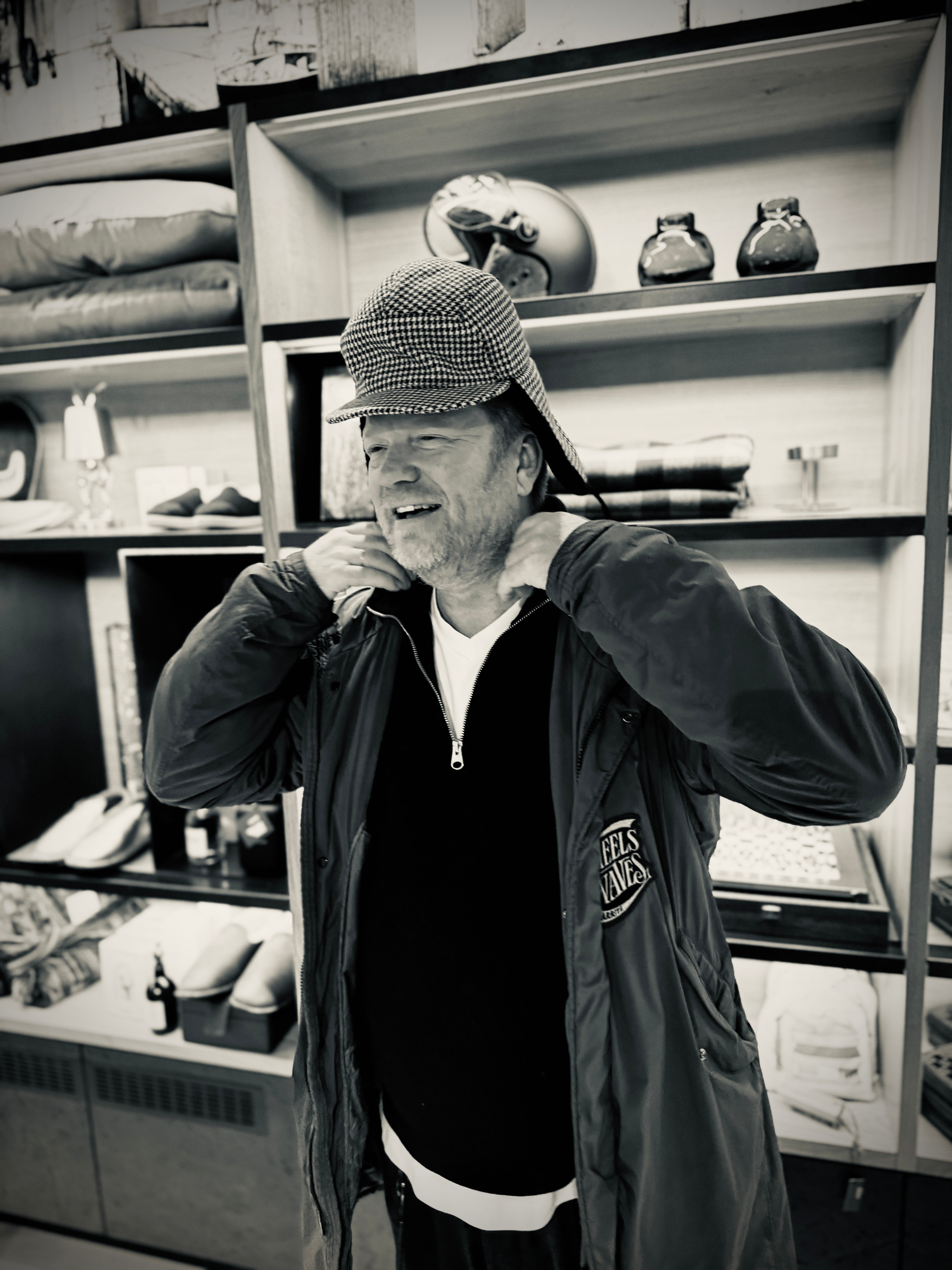 Alex Rivera, lachend mit Schiebermütze und Wheels-&-Waves-Jacke in einem Concept Store – Merchant & Friends Friend