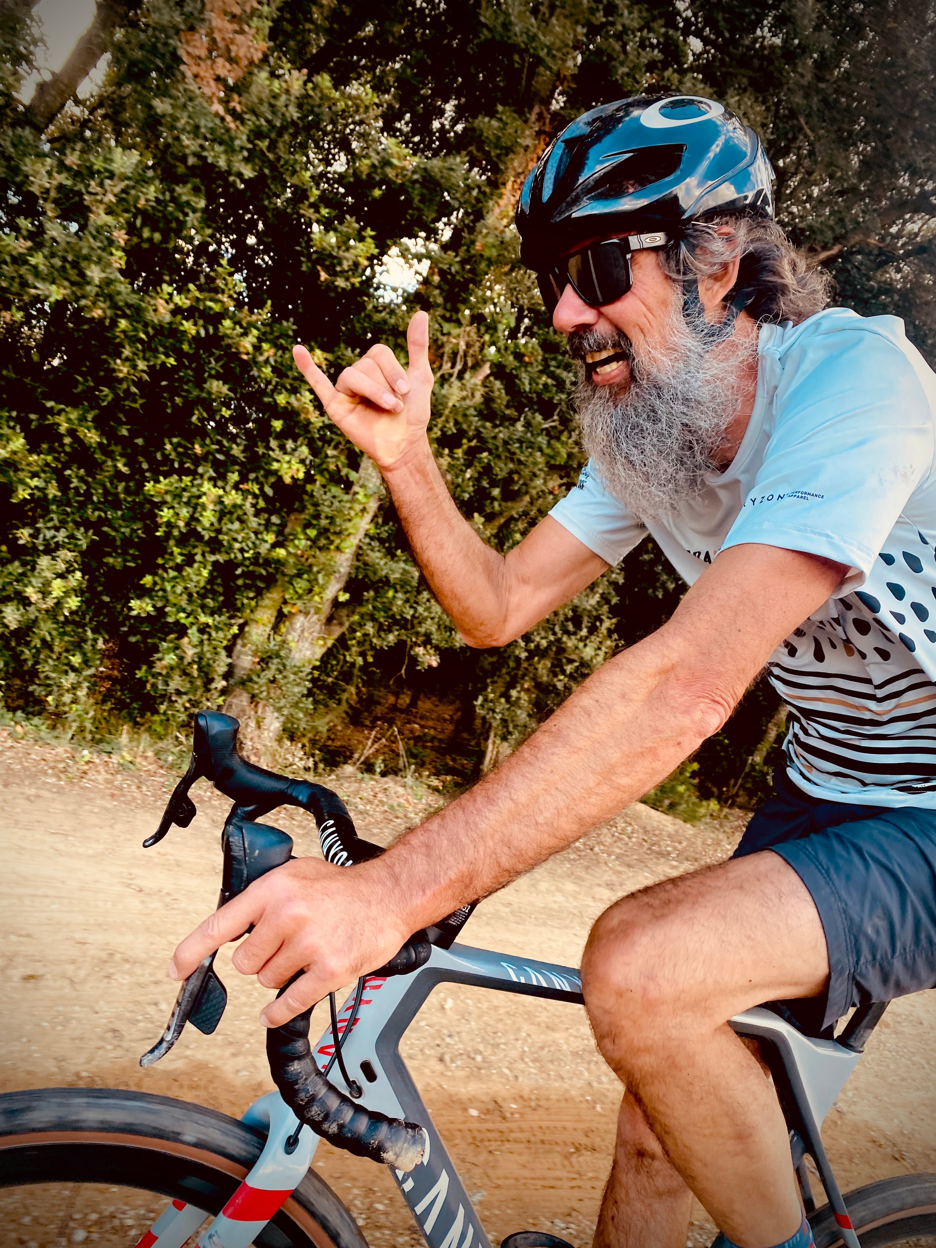 Chris Walter mit langem grauen Bart auf dem Gravelbike, lachend mit Shaka-Geste auf einem Waldweg – Merchant & Friends Friend