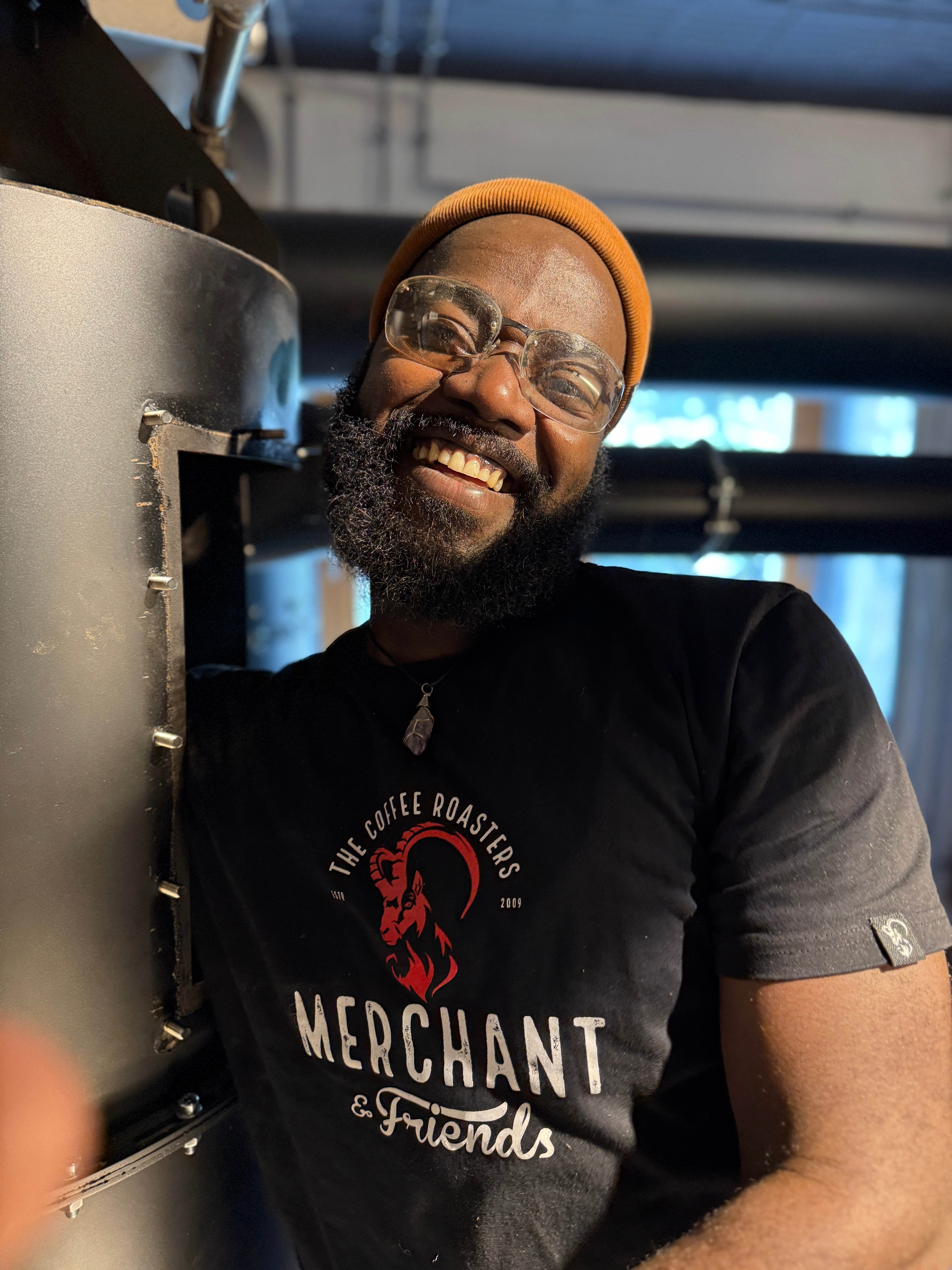 José im Merchant-&-Friends-Shirt, breit lächelnd an der Kaffeerösttrommel – Merchant & Friends Friend